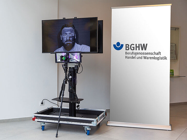 UV-Kamera auf Stativ mit Monitoren, UV-Strahlern und einem Unterbau in aufgebautem Zustand, daneben ein Roll up mit der Aufschrift BGHW 