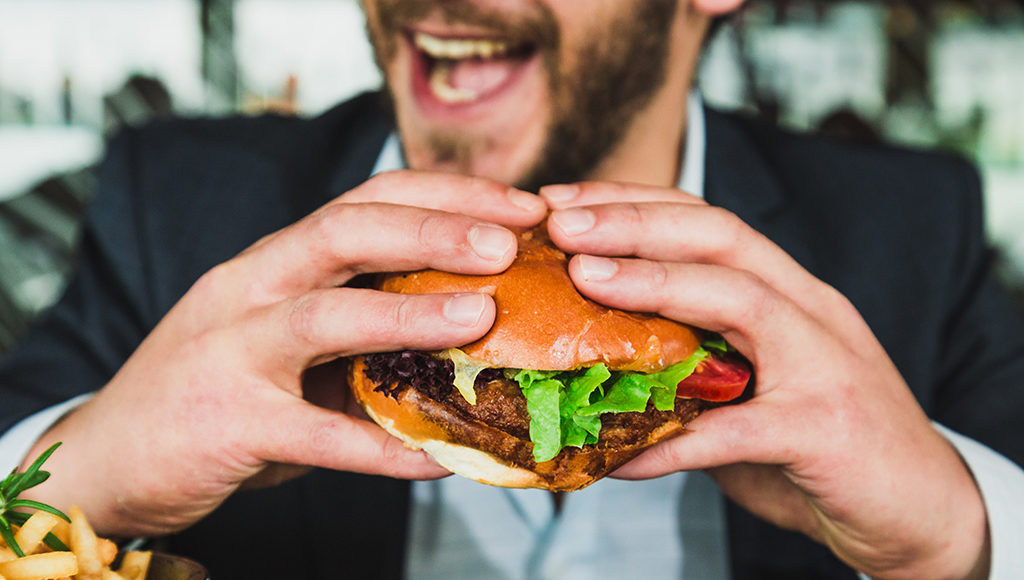 Zu sehen ist ein Hamburger im Vordergrund und ein lachender Mann im Hintergrund